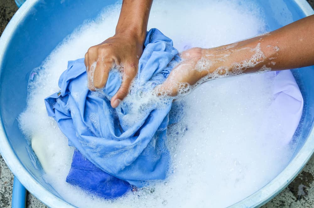 Close up of hands washing a blue clothing item in a blue bucket with soap and water
