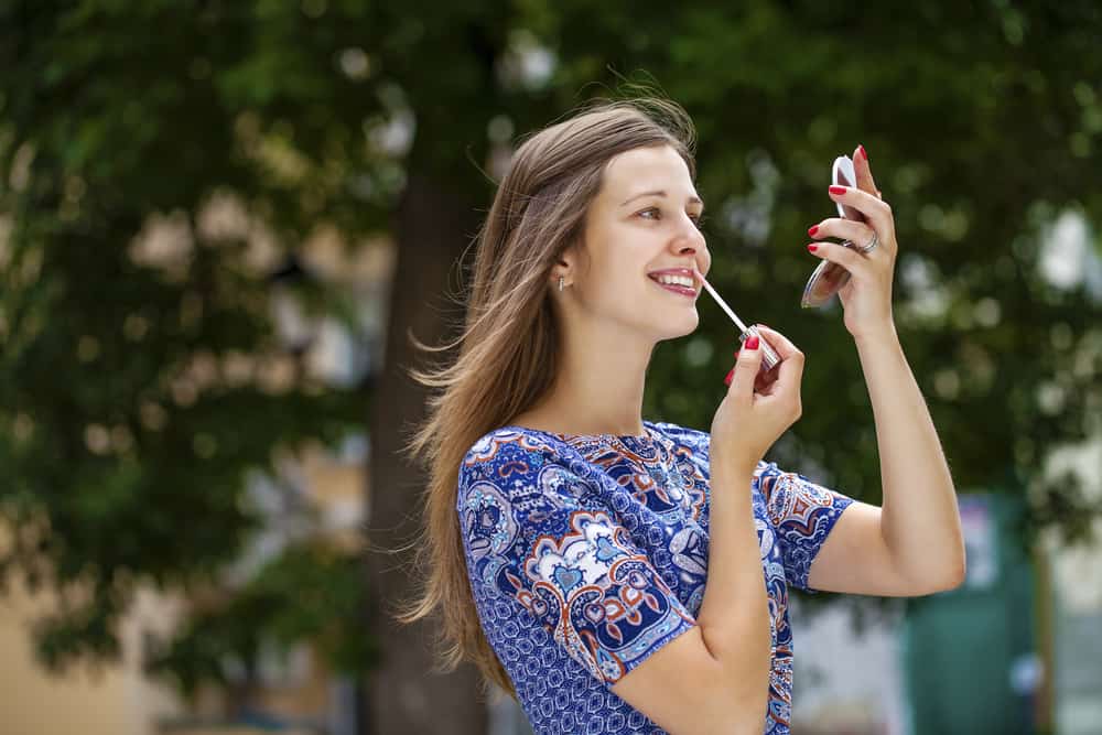 pretty woman applying lip tint with pocket mirror outside
