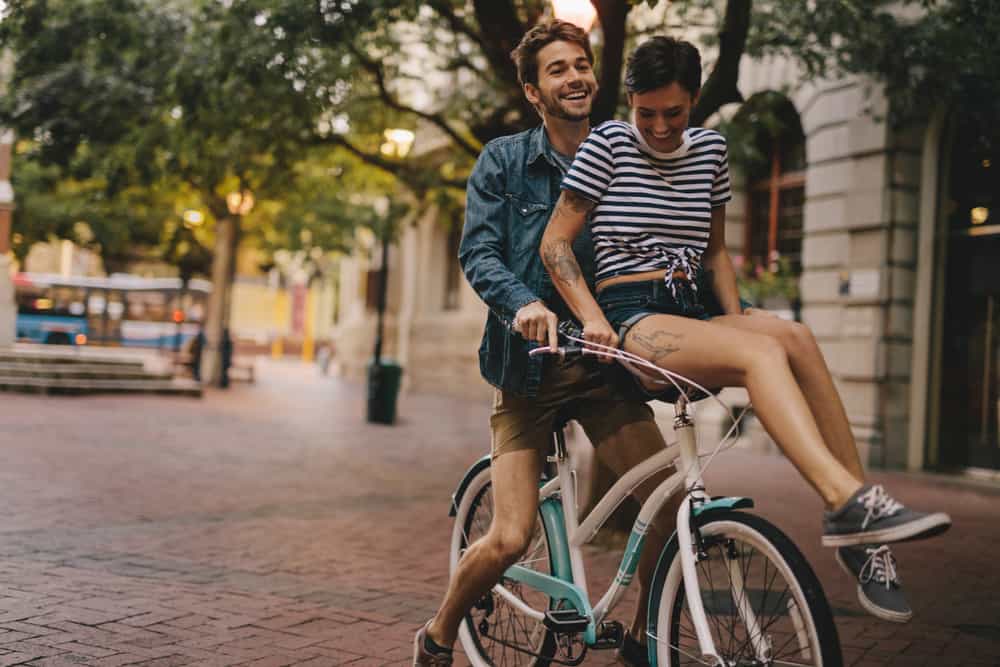 a woman sits on the handlebars of a bicycle in front of a smiling man