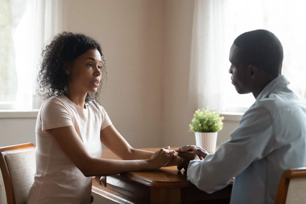 an african-american couple sits holding hands while the woman speaks with a serious expression